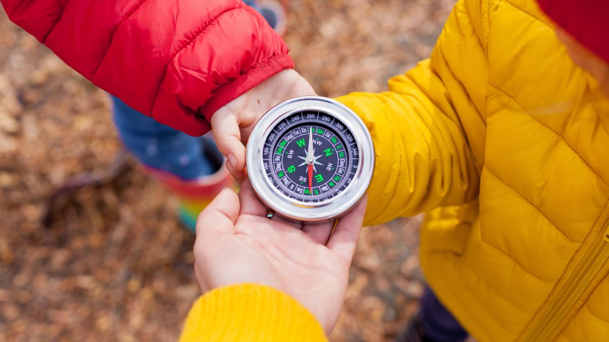 Close-up of three children's hands holding a compass. Children wear red and yellow winter jackets.