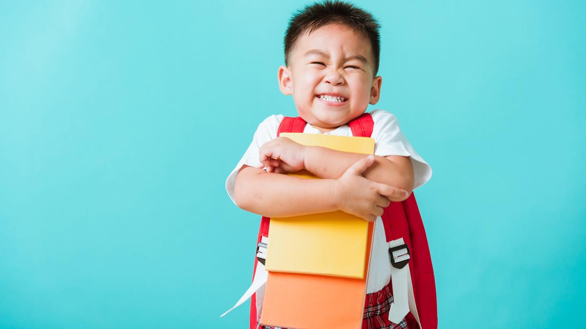 Preschool boy wearning red backpack hugs books and grins.