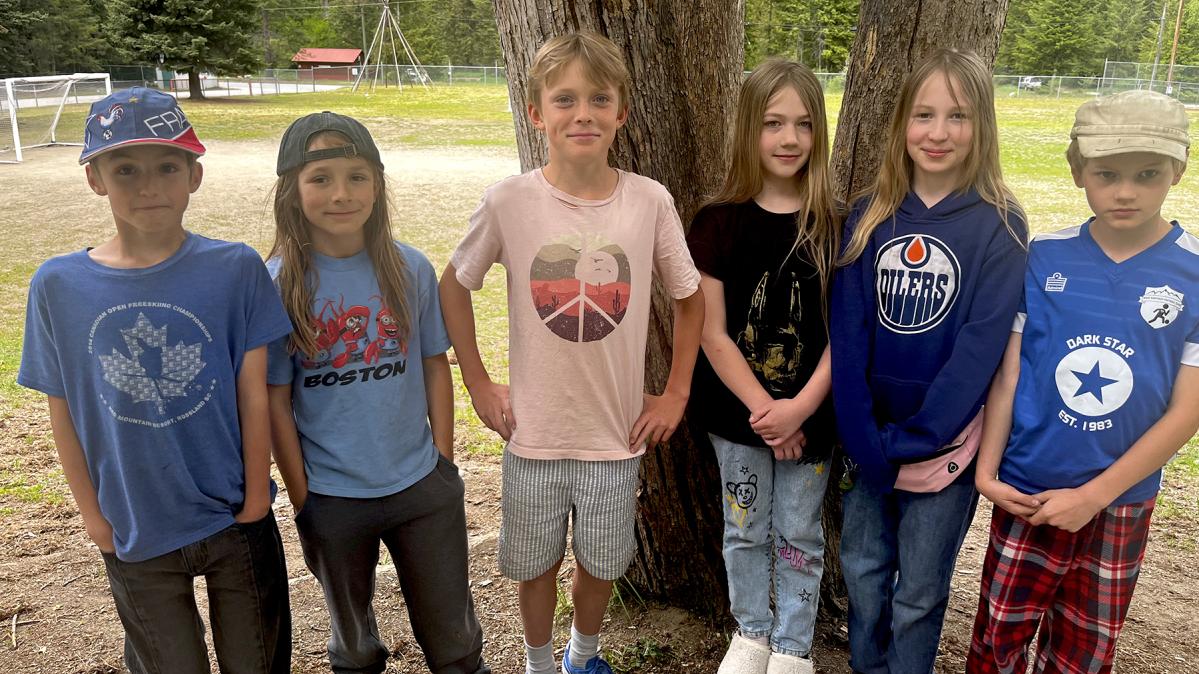 Six children stand outside in front of two trees with a school playground behind them.