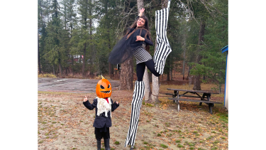 A child with a pumpkin on their head and an adult woman on stilts covered in striped pants strike a pose in a forest.