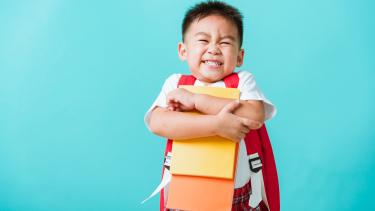 Preschool boy wearning red backpack hugs books and grins.