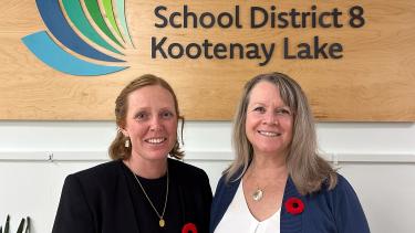 A woman with red hair in a black suit stands beside a woman with blonde, shoulder-length hair in a blue sweater and white shirt, in front of a wooden sign indoors.
