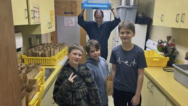 Three boys in a commercial kitchen with an adult standing behind them holding a dish tray